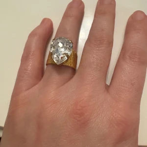 Statementring Carolina Svedbom guld - Unik statementring från Carolina Svedbom med bred, hamrad guldfärgad ring och stor droppformad klar kristall. Ringen är öppen baktill och har en lyxig, glittrig känsla som verkligen drar blickarna till sig.