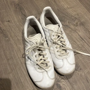 Vita Adidas Gazelle sneakers - Säljer ett par klassiska vita Adidas Gazelle sneakers med de ikoniska tre ränderna på sidan och guldfärgad Gazelle-text. Skorna har snörning, platt sula och rund tå. Tillverkade i skinn med detaljer i syntet. Perfekt för dig som gillar retrostil och enkel design.