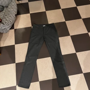 Grå kostymbyxor från Grunt, stl 27 - Snygga ljusgrå kostymbyxor från Grunt, modell Dude Pant. Byxorna har raka ben, klassiska fickor fram och bak samt hällor för bälte. Perfekt för dig som gillar stilrena och enkla byxor med modern passform.