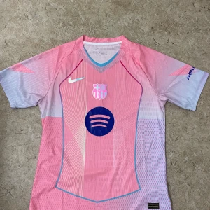 FC Barcelona rosa matchtröja Nike - Snygg FC Barcelona fotbollströja från Nike i rosa och ljuslila med korta ärmar. Tröjan har klubbmärke, Nike-logga och Spotify som sponsor framtill samt UNHCR-logga på ryggen. Tillverkad i lätt och ventilerande polyester med grafiskt mönster och V-ringad hals.
