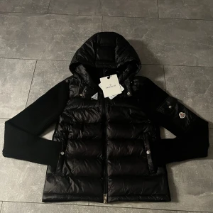Moncler cardigan svart - Säljer en svart Moncler dunjacka med stickade ärmar och huva. Jackan har dragkedja framtill, fickor med tryckknappar och en ikonisk Moncler-logga på ärmen. Materialmix med dunfyllning och ribbstickade detaljer ger en riktigt snygg look.