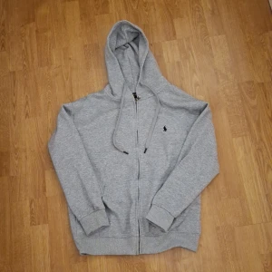 Grå hoodie från Polo Ralph Lauren - Snygg grå hoodie från Polo Ralph Lauren med dragkedja och huva med snörning. Klassisk broderad logga på bröstet och ribbade muddar vid ärmar och nederkant. Perfekt för chill dagar och enkel att matcha med jeans eller joggers. Skriv om ni har frågor.