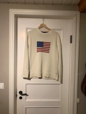 Vit stickad tröja från GANT - Vit stickad tröja från GANT med amerikansk flagga broderad på bröstet och GANT New York 1949-tryck på ärmen. Tröjan har rund hals, ribbade muddar och är långärmad. Perfekt för dig som gillar klassisk och clean stil.