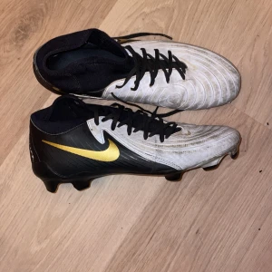 Nike phantom fotbollsskor svart/vit/guld - Snygga fotbollsskor från Nike med svart och vit ovandel, gul swoosh på sidan och snörning. Skorna har en strömlinjeformad design med rund tå och platt sula med dobbar för bra grepp på planen. Perfekt för dig som vill sticka ut på matchen.