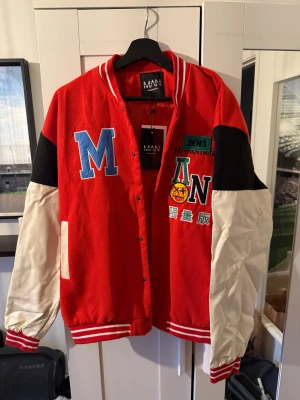 Röd bomberjacka MAN Limited Edition - Limited Edition bomberjacka från MAN med röd kropp, vita ärmar och svarta axelpartier. Jackan har broderade och tryckta detaljer fram och bak, ribbade muddar med vita ränder och knappar framtill. Snygga patchar och text ger jackan en unik streetstil.