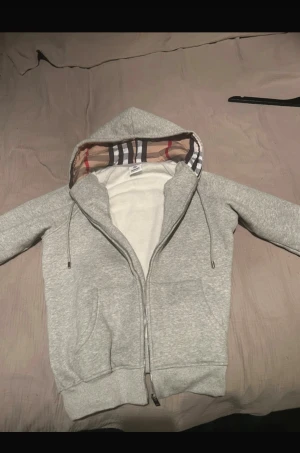 Grå hoodie från Burberry med dragkedja - Säljer en grå hoodie från Burberry i storlek M. Tröjan har klassisk Burberry-rutig insida på huvan, snörning och dragkedja framtill. Mjuk bomullskänsla, två fickor och ribbade muddar. Perfekt för dig som gillar stilrena och lyxiga streetwear-plagg.