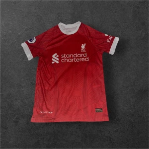 Liverpool röd fotbollströja Nike - Säljer en officiell Liverpool FC fotbollströja från Nike i rött med vita detaljer vid krage och ärmslut. Tröjan har korta ärmar, klubbmärke och sponsortryck på bröstet samt Premier League-märke på ärmen. Tillverkad i lätt, ventilerande material.