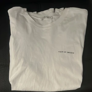 Vit t-shirt från Tiger of Sweden - Vit t-shirt från Tiger of Sweden med minimalistisk svart logga på bröstet. Klassisk rund halsringning och stilren design, perfekt för en clean och enkel look. Strlk M. Skriv för frågor