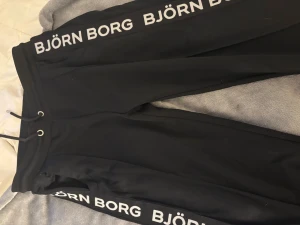 Svarta mjukisbyxor från Björn Borg - Svarta mjukisbyxor från Björn Borg med vit logga längs båda benen. Byxorna har snörning i midjan och är tillverkade i ett mjukt material som passar perfekt för chill eller träning. Snygg och sportig look med normal passform.