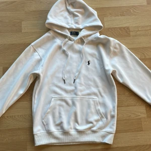 Vit hoodie från Polo Ralph Lauren - Vit hoodie från Polo Ralph Lauren med klassisk broderad logga på bröstet. Tröjan har huva med snörning, stor magficka och ribbade muddar. Perfekt för chill dagar och enkel att matcha med jeans eller joggers.