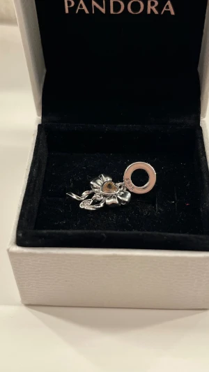 Pandora silverberlock med rosett och hjärta - Snygg berlock från Pandora i silverfärg med en detaljrik rosett och ett litet hjärta i mitten. Perfekt att hänga på ditt armband eller halsband för att ge en personlig touch. Hjärtdetaljen har en varm ton som sticker ut mot det blanka silvret.