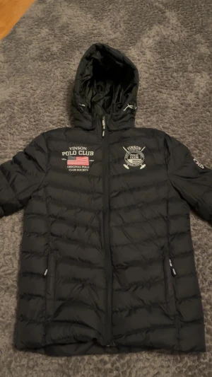 Svart pufferjacka från Vinson Polo Club - Svart pufferjacka från Vinson Polo Club med broderade loggor och amerikansk flagga på bröstet och ärmen. Jackan har huva med snörning, två sidofickor med dragkedja och quiltad design. Perfekt för kalla dagar med stilren look.