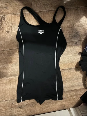 Svart baddräkt från Arena - Sportig svart baddräkt från Arena med vita linjer längs sidorna och klassisk logga framtill. Modellen har breda axelband och är designad för simning och träning. Materialet är syntet och sitter tight mot kroppen för bästa rörelsefrihet i vattnet. Har lite mer av en shorts underdel