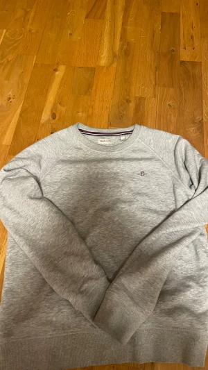 Grå sweatshirt från GANT - Grå sweatshirt från GANT med rund hals och broderad logga på bröstet. Tröjan har ribbade muddar vid ärmslut och nederkant samt en diskret röd, vit och marinblå rand på insidan av halsen. Perfekt för chill dagar och passar fint till gråa, mörkblåa ock svarha jeans.