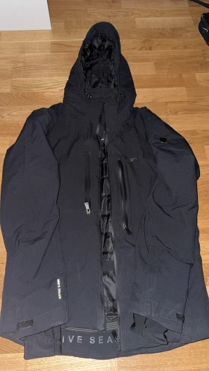 Svart pufferjacka från Five Seasons - Svart pufferjacka från Five Seasons med huva och flera dragkedjeförsedda fickor. Jackan har quiltad insida och justerbara ärmslut med kardborre. Perfekt för kalla dagar med sin tjocka och isolerande design. Snygg och funktionell för dig som gillar stil och värme.