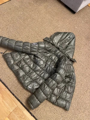 Grön jacka Hampton Republic - Snygg grön jacka från Hampton Republic med lila foder och huva. Jackan har quiltad design, två dragkedjeförsedda fickor och coolt badge på bröstet. Lila detaljer och logga på framsidan.