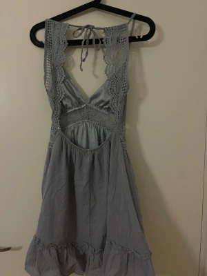 Blå kort klänning med spetsdetaljer - Supersöt blå kort klänning med spetsdetaljer längs axelbanden och midjan. Klänningen har v-ringning, knytning i nacken och volangkant nertill. Perfekt för varma dagar och har en luftig passform med fina virkade detaljer.