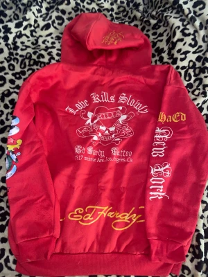 Röd Ed Hardy zip hoodie - Snygg röd zip hoodie från Ed Hardy med ikoniska tattoo-inspirerade tryck i vitt, gult och fler färger. Jackan har huva, dragkedja och fickor fram. Detaljer som döskalle, texter och motiv på både bröst och ärmar ger en unik streetstil.