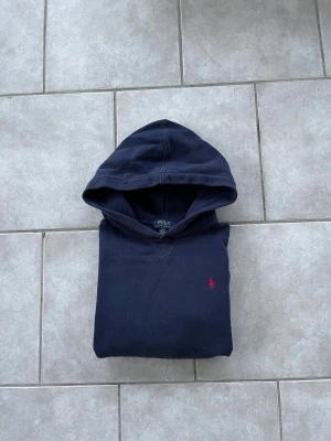 Mörkblå hoodie från Polo Ralph Lauren - Snygg mörkblå hoodie från Polo Ralph Lauren med klassisk röd broderad logga på bröstet. Tröjan har huva, långa ärmar och ribbade muddar. Perfekt för dig som gillar stilrena och tidlösa plagg. 