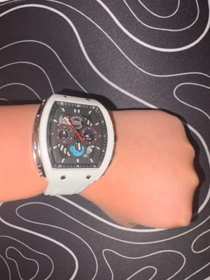 En snygg och tuff QFNS klocka! - En stilren och robust klocka i vit finish med tonad glasruta och flera dekorativa subdials för en exklusiv look. Svart urtavla med markerade index och röda detaljer ger klockan ett sportigt, lyxigt uttryck. Bekväm att bära och passar både vardag och fest. Fint skick! Tveka inte att skriva!😄⌚️