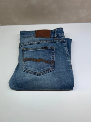 Blå Nudie Jeans - Snygga blå jeans från Nudie Jeans med klassisk femficksdesign och bruna sömmar på bakfickorna. Jeansen har normal passform och är tillverkade i mjukt bomullsjeansmaterial. Perfekt för dig som gillar stilrena och tidlösa jeans. WAIST 29 lengh 32