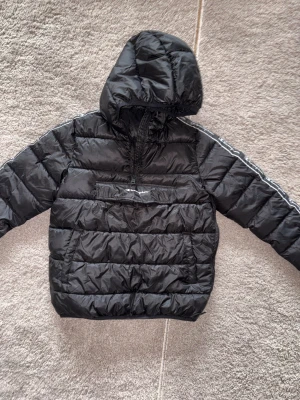 Svart pufferjacka från Champion  - PRIS KAN DISKUTERAS || Svart pufferjacka från Champion i storlek XL. Jackan har huva, halv dragkedja och en stor ficka framtill med Champion-logga. Quiltad design och sportiga detaljer längs ärmarna. Perfekt för kalla dagar och har en loose passform.