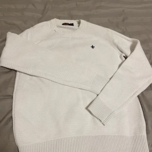 Vit stickad tröja från Morris - Säljer en stilren vit stickad tröja från Morris med ett diskret broderat emblem på bröstet. Tröjan har långa ärmar och ribbade muddar vid ärmslut och nederkant. Perfekt för en klassisk look.