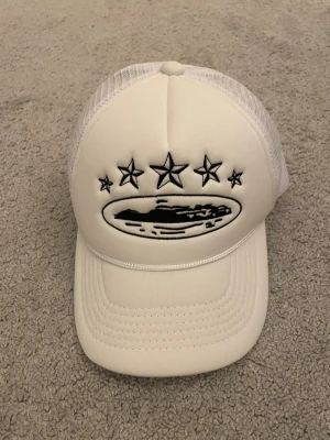 Vit Corteiz truckerkeps med stjärnor - Snygg vit truckerkeps från Corteiz med svart broderade stjärnor framtill och på sidan. Kepsen har mesh på sidorna och bak, samt broderad Corteiz-logga baktill. Framsidan pryds av ett ovalt motiv och stjärnor. Perfekt för dig som gillar streetwear och vill sticka ut.