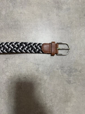 Svartvitt flätat bälte med skinn - Snyggt flätat bälte i svart, vitt och grått med bruna skinn-detaljer vid spännet och änden. Bältet har ett klassiskt metallspänne och passar perfekt till jeans eller chinos för en stilren look.