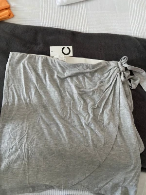 Kjol - Snygg grå kort kjol från Cubus i mjuk bomullsmix. Kjolen har en draperad effekt och knytning i sidan för en cool och avslappnad look. Perfekt för dig som gillar enkel och stilren design med en twist. Helt ny med prislappen på.