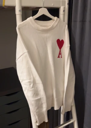 Vit stickad Ami Paris tröja - Vit stickad tröja från Ami Paris med röd hjärtlogga och bokstaven A på bröstet. Tröjan har rund hals, ribbade muddar och lång ärm. Perfekt för dig som gillar clean stil med en twist. Materialet är mjukt och håller värmen bra.