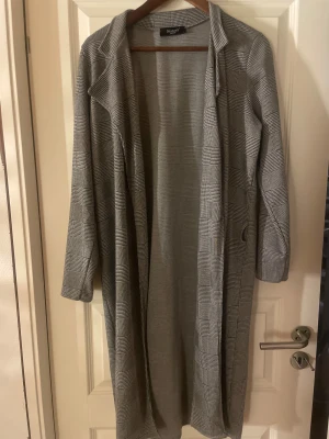 Grå rutig lång kappa från Sisters point  - Snygg lång kappa från Sisters i grått med rutigt mönster. Klassisk krage och långärmad modell med avslappnad passform. Perfekt för dig som gillar stilrena och tidlösa plagg. Materialet känns mjukt och lätt, vilket gör den enkel att bära under höst och vår.