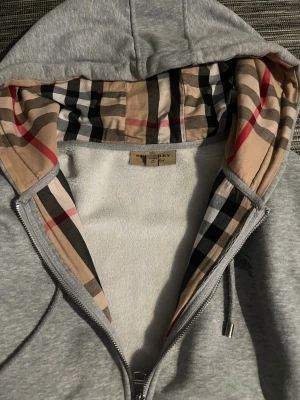 Grå Burberry hoodie med rutig huva - Snygg grå hoodie från Burberry med dragkedja och klassisk rutig insida i huvan. Tröjan har broderad logga på bröstet, snörning i huvan och två fickor framtill. Perfekt för dig som gillar stilrena detaljer och ikoniskt mönster. Sår storlek xl men är Mellan M och L, passar båda bra