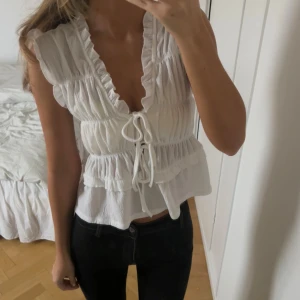 Vit blus med volanger och snörning - Säljer en söt vit blus med volanger och djup v-ringning. Toppen har snörning framtill och är croppad med flera lager volanger. Materialet är lätt och luftigt, perfekt för varma dagar. Ärmlös design med romantiska detaljer som ger en fräsch och trendig look.