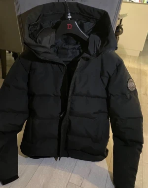 Svart pufferjacka från Canada Goose - Säljer en svart pufferjacka från Canada Goose med huva och snyggt emblem på ärmen. Jackan har dragkedja framtill och är fodrad för extra värme. Perfekt för kalla dagar och har en stilren look som funkar till det mesta.