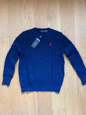 Marinblå kabelstickad tröja Polo Ralph Lauren - Kabelstickad marinblå tröja från Polo Ralph Lauren med klassisk rund hals och röd broderad logga på bröstet. Tröjan har långa ärmar och ribbade muddar vid ärmslut och nederkant. Perfekt för dig som gillar stilren och tidlös design. Pris kan diskuteras 