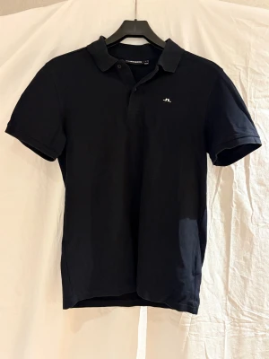 Svart pikétröja från J.Lindeberg - Klassisk svart pikétröja från J.Lindeberg i storlek S. Tröjan har korta ärmar, krage med två knappar och en liten vit logga broderad på bröstet. Tillverkad i mjuk bomull som andas och passar perfekt till en clean och stilren look.