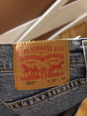 Levi's 502 blå jeans W30 L32 - Klassiska Levi's 502 jeans i blå denim med raka ben och normal passform. Jeansen har fem fickor, dragkedjegylf och tydliga gula sömmar. Perfekt för dig som gillar en tidlös look med en touch av vintage.