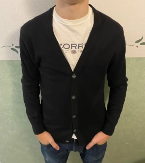 Svart kofta från Christian Berg - Svart kofta från Christian Berg i storlek M. Klassisk v-ringad modell med knappar framtill och långa ärmar. Mjuk och lätt stickad känsla, perfekt att slänga över en t-shirt eller skjorta. Enkel och stilren design som funkar till jeans eller chinos.