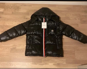 Svart Moncler pufferjacka med huva - Svart pufferjacka från Moncler med glansig finish, huva och ikonisk logga på bröstfickan. Jackan har en rödvit dragkedja framtill och praktiska fickor med dragkedja. Perfekt för kalla dagar och har en riktigt clean look. Själv har jag storlek s men passar mig lite gran bara att den är lite tjock på mig men skulle rekommendera om du har m i storlek att köpa den då