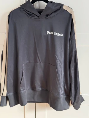 grå hoodie med beige ränder - Snygg grå hoodie från Palm Angels med beige ränder längs ärmarna och vit logga på bröstet. Tröjan har huva, stor magficka och långärmad passform. Materialet är mjukt och har en lätt glansig finish som ger en cool streetkänsla.