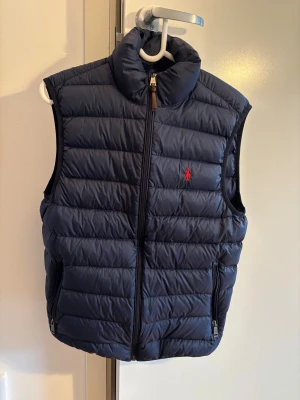 Marinblå dunväst från Polo Ralph Lauren - Snygg marinblå dunväst från Polo Ralph Lauren med röd logga på bröstet. Västen har quiltad design, hög krage och två praktiska fickor med dragkedja. Bra skick, Storlek S. Har du några frågar så hör gärna av dig. 