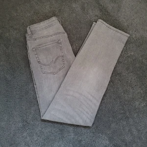 Grå jeans från Jack & Jones - Säljer ett par grå jeans från Jack & Jones, modell Regular Clark. Jeansen har bälteshällor, knapp och dragkedja framtill. Perfekta för dig som gillar en enkel och stilren look.