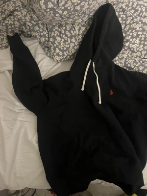 Svart hoodie från Polo Ralph Lauren - Svart hoodie från Polo Ralph Lauren med klassisk röd logga på bröstet. Tröjan har huva med vita snören och en stor ficka framtill. Mjuk och skön, perfekt för chill dagar. Passar dig som gillar enkel och stilren streetwear.