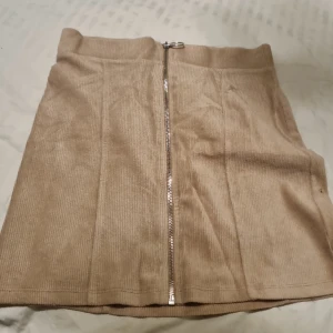 Beige manchesterkjol från Gina Tricot - Säljer en beige kortkjol i manchester från Gina Tricot. Kjolen har en rak passform och en snygg silverfärgad dragkedja framtill som detalj. Perfekt att matcha med stickad tröja eller t-shirt för en trendig look.