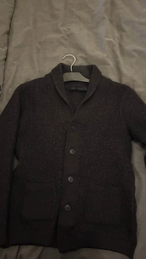 Svart kofta från Riley - Säljer en svart stickad kofta från Riley med klassisk krage och fyra knappar framtill. Koftan har två praktiska fickor och långärmad design, perfekt för lager-på-lager under kyliga dagar. Materialet är mjukt och ger en chill vibe.