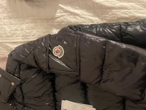 Svart Moncler pufferjacka M - Svart pufferjacka från Moncler med glansig finish och klassisk logga på ärmen. Jackan har hög krage, dragkedja framtill och två sidofickor med dragkedja. Insidan har en cool comic patch. Perfekt för dig som vill ha en snygg och varm jacka med street vibe.