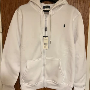 Vit hoodie från Polo Ralph Lauren - Vit hoodie från Polo Ralph Lauren med dragkedja, snörning i huvan och klassisk svart logobrodyr på bröstet. Tröjan har två stora fickor framtill och ribbade muddar vid ärmslut och nederkant. Perfekt för chill dagar och enkel att matcha med jeans.