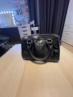 Väska som är inspirerad av zadig zoltaire - Cool svart handväska i skinn med silverfärgade detaljer och ett snyggt emblem med vingar på framsidan. Väskan har både handtag och en avtagbar axelrem, samt dragkedjor på sidorna för extra edge. Perfekt storlek för det viktigaste och ger en grym vibe till din outfit. Pris kan diskuteras!