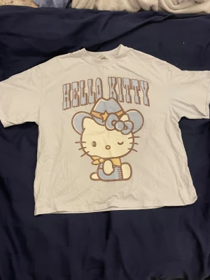 Hello Kitty cowboy t-shirt från H&M - Vit t-shirt från H&M med stor Hello Kitty-tryck där hon har cowboyhatt och rosett i ljusblått och beige. T-shirten har korta ärmar och en loose passform, perfekt för dig som gillar söta och roliga motiv. Materialet är mjuk bomull.